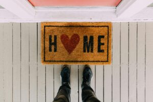 Person stands at the entrance to a home; we see his feet and a doormat that says "Home" with the o shaped as a heart.
